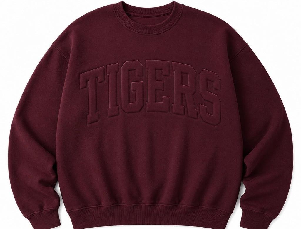 A crewneck sweatshirt in a deep burgundy color, featuring the word 'TIGERS' embossed across the chest in matching dark lettering.