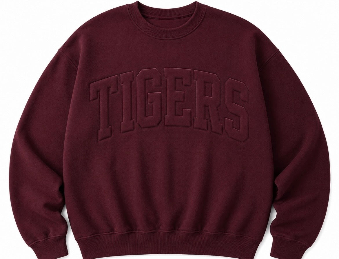 A crewneck sweatshirt in a deep burgundy color, featuring the word 'TIGERS' embossed across the chest in matching dark lettering.